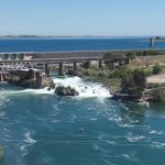 Wide-angle look upstream at the Snake River toward American Falls Dam. Water in the foreground is bright blue, Some whitewater is spilling from the dam, and the sky above is blue.