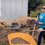 Three smiling Idaho Power volunteers in teal shirts using shovels to fill a yellow wheelbarrow.