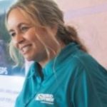 Blonde woman wearing a teal Idaho Power shirt standing in front of a large map talks with another blonde woman in a nondescript meeting room.