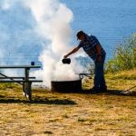 Permalink to Stay Safe and Prevent Power Outages this Fourth Man pouring water from a tea kettle over a campfire in an established campfire ring in front of a lake with mountains in the background