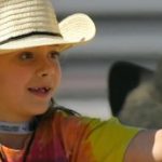 A smiling young girl wearing a straw hat and tie-dye t-shirt giving a high-five to another cowboy.